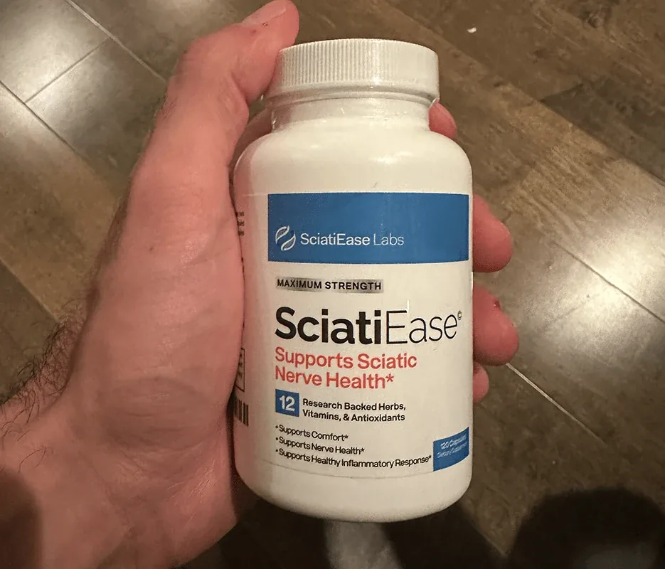 A hand holding a white SciatiEase bottle over a wood floor: SciatiEase Labs band, MAXIMUM STRENGTH, Supports Sciatic Nerve Health, 120 Capsules
